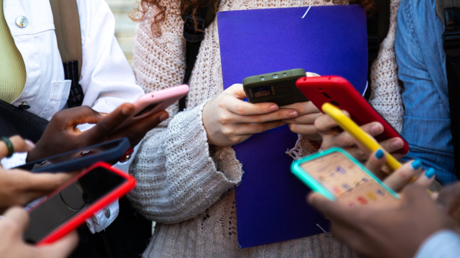 A group of young people stand huddled in a circle using smartphones. 