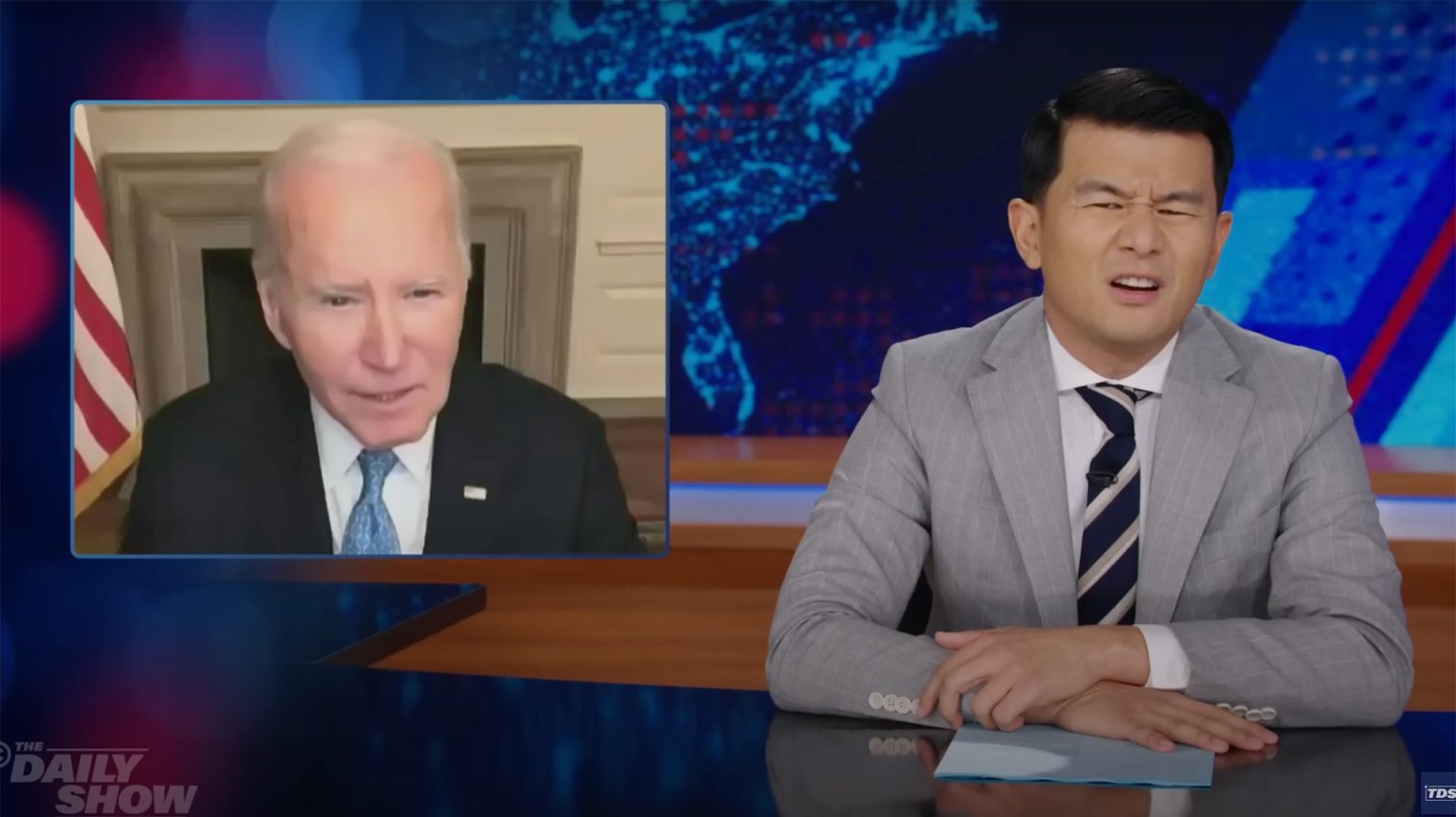 A man in a suit sits behind a talk show desk looking confused. In the top-left is an image of another man on a video call.