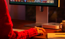 a close-up of a mac mini underneath an apple studio display next to a mug, a notebook, and a woman's hand on a mouse in dim, moody lighting