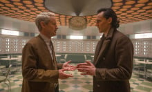 Two men face each other holding out their hands, standing in a retro looking room.