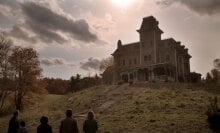 A group of people stand at the foot of a hill, looking up at a large house.