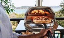 a pizza going into an ooni oven with a fire lit. the background is a beautiful body of water and mountain in the distance.