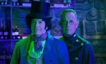 Two men in costume stand in a green lit room, staring at the camera.