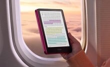 a person holds the new kindle colorsoft signature edition in front of an airplane window that shows a sunset sky with puffy clouds below