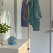 a twilight blue echo dot sitting on a white console table next to a potted plant and a wood tray filled with keys, sunglasses, and a wallet. the table is next to a door and a coat rack holding several jackets