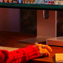 a close-up of a mac mini underneath an apple studio display next to a mug, a notebook, and a woman's hand on a mouse in dim, moody lighting