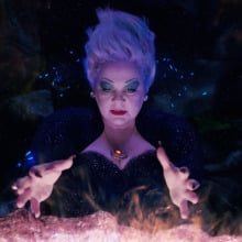 A sea witch wearing a bedazzled black dress stands in front of a glowing cauldron. 