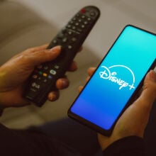 Hands holding a TV remote and a smartphone showing the Disney Plus logo