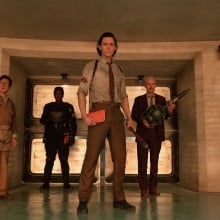 A man in a mechanic suit, a woman in black armor, and two men in suits stand in a control room.