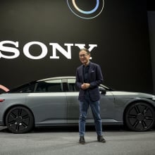 An asian man in a suit stands in front of a car. In the background is a giant screen with the words Sony displayed