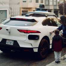 A woman and her child stand outside a white Waymo driverless vehicle, seen from the back. The woman's hand is on the car door's handle and it appears she is about to open the door.