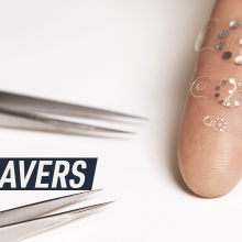 A white background photograph shows a couple of medical tweezers next to three of the tiny stickers placed on an index finger for scale.