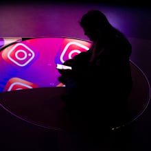 A person sitting in a dark room on their phone. A pattern showing the Instagram logo is reflected in mirrors below them. 