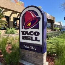 Taco Bell drive-thru open for business with lush green plants and clear sky in a residential neighborhood