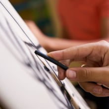 A close-up of a person's hand holding charcoal as they draw on an easel at an art class. 