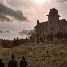 A group of people stand at the foot of a hill, looking up at a large house.