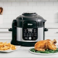 a ninja foodi air fryer and pressure cooker combo sits on a kitchen counter with several dishes of food in front of it including a roast chicken, french fries, and brussel sprouts