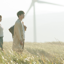 A still from 'Beyond Goodbye' of a man and woman in a field, with wind turbines in the background.