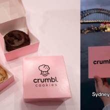 A composite of two images. On the left, three boxes of Crumbl Cookies, two of them open showing the cookies inside. On the right, a screenshot of a video from YouTube channel CrumblCookieAus, showing hands holding up a Crumbl Cookie and box in front of the Sydney Harbour Bridge. Text on the screenshot reads, "Sydney we are here!"