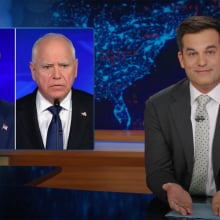 A man in a suit sits behind a talk show desk. In the top-left is a side-by-side image of two other men in suits.