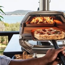 a pizza going into an ooni oven with a fire lit. the background is a beautiful body of water and mountain in the distance.