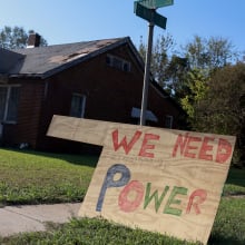 A wooden sign that reads "We need power" on the lawn outside of a home.