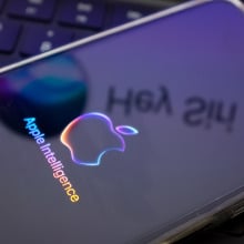 The Apple Intelligence logo is being displayed on a smartphone, with the Apple Siri logo in the background