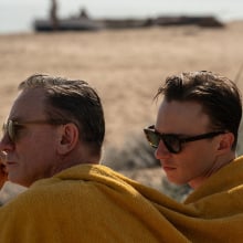 Daniel Craig and Drew Starkey sit huddled under one yellow towel on the beach in a still from the film "Queer."