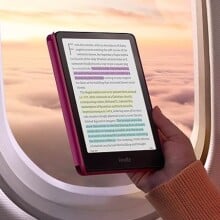 a person holds the new kindle colorsoft signature edition in front of an airplane window that shows a sunset sky with puffy clouds below