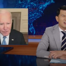 A man in a suit sits behind a talk show desk looking confused. In the top-left is an image of another man on a video call.