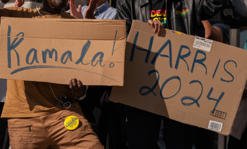 Two people each hold up a cardboard sign. One reads, "Kamala!" The other reads "Harris 2024".