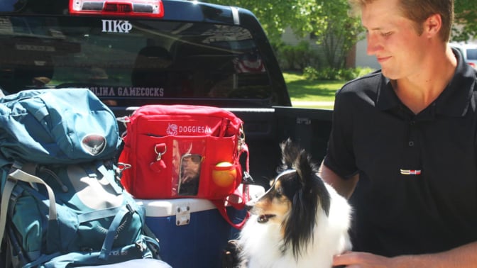 Person sitting with dog in back of truck with red doggiesak bag on top of cooler and blue bags next to them