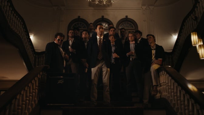 A group of frat brothers in suits stand on a grand staircase in a frat house.