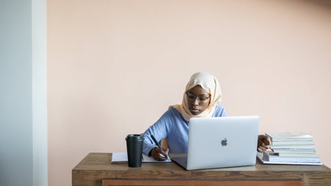 Woman sitting in front of an open laptop and writing notes with books and a cup of coffee nearby.