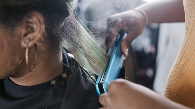 A close up of a black hairdresser straightening a client's hair with some steam rising from the heat.