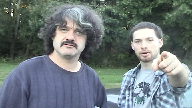 Two men stand outside outside, one is pointing at the camera.