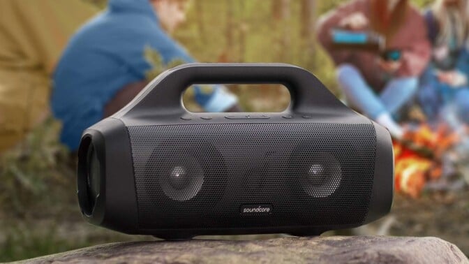 a soundcore anker motion boom speaker sits on a rock outside with people sitting behind it