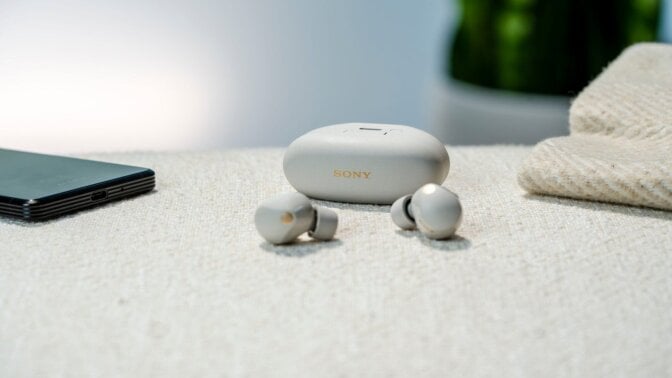 a pair of sony WF-1000XM5 wireless earbuds sit on a table with a cell phone off to the left