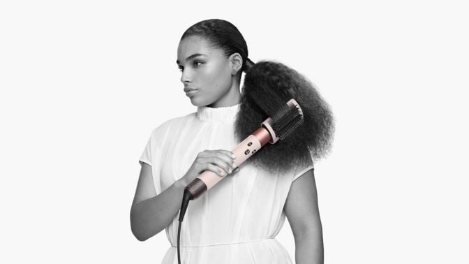 a person stands against a white background while using the dyson airwrap complete long hair styling tool with a brush attachment