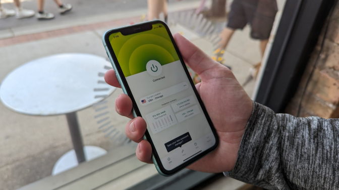 a close-up of a man holding an iphone displaying the expressvpn app in front of a window