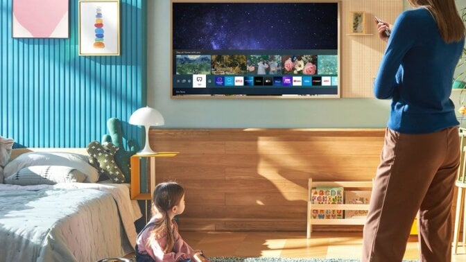 Parent pointing remote at Samsung The Frame TV hanging on wall and child sitting on floor near bedroom furniture in peripherals