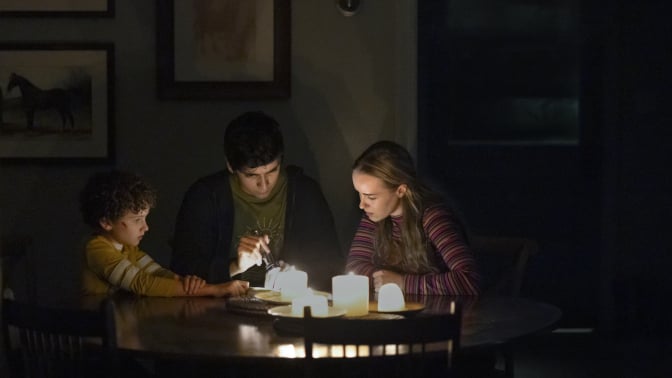 A family sit in their home in the dark, at the dining table lit by candles.