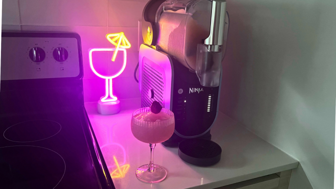 Glass of frosé on countertop beside Ninja Slushi machine with neon cocktail light in background