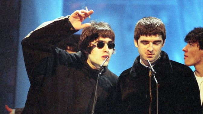 liam and noel gallagher on stage at an award ceremony. liam is gesturing with a cigarette in his hand, noel is looking downward.