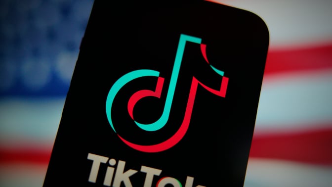 An phone displaying the TikTok logo in front of a blurry American flag. 