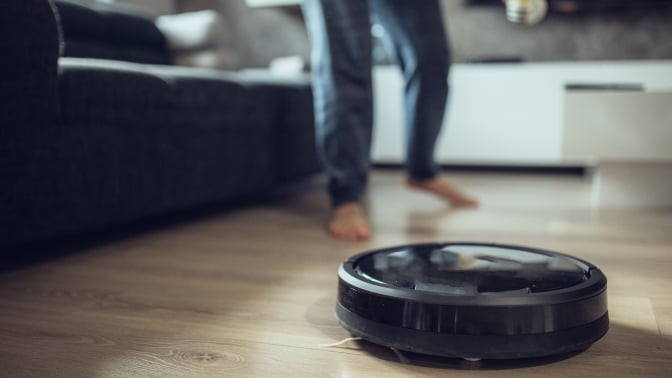 A robot vacuum on a wooden floor. A person's legs are seen running behind it.