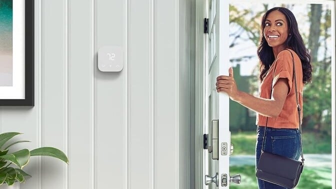 a person leaves through the front door of a home while looking back into the home and smiling. there's an amazon smart thermostat affixed to an interior wall inside the home