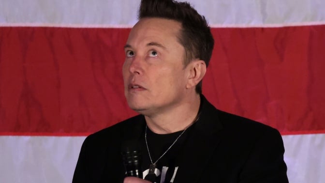 Elon Musk stares upwards in front of a US flag. 