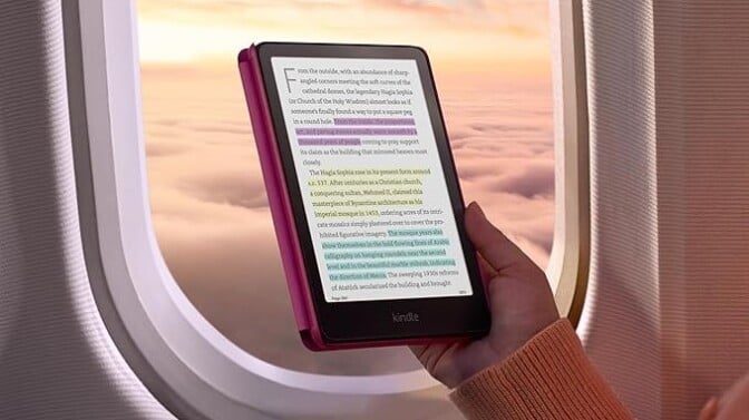 a person holds the new kindle colorsoft signature edition in front of an airplane window that shows a sunset sky with puffy clouds below