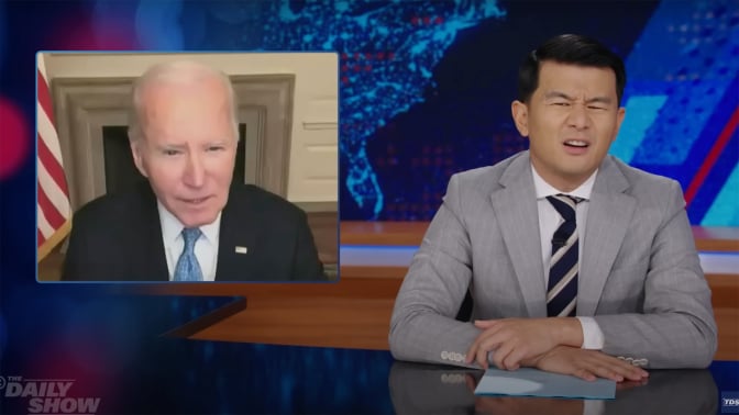 A man in a suit sits behind a talk show desk looking confused. In the top-left is an image of another man on a video call.